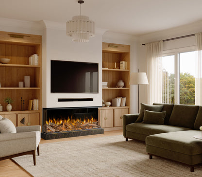 A modern living room featuring a built-in electric fireplace with a black frame and glowing logs, flanked by light wood built-in shelves. A green chaise lounge, a gray armchair, and a textured beige rug are arranged in front of the fireplace under a white tiered chandelier.